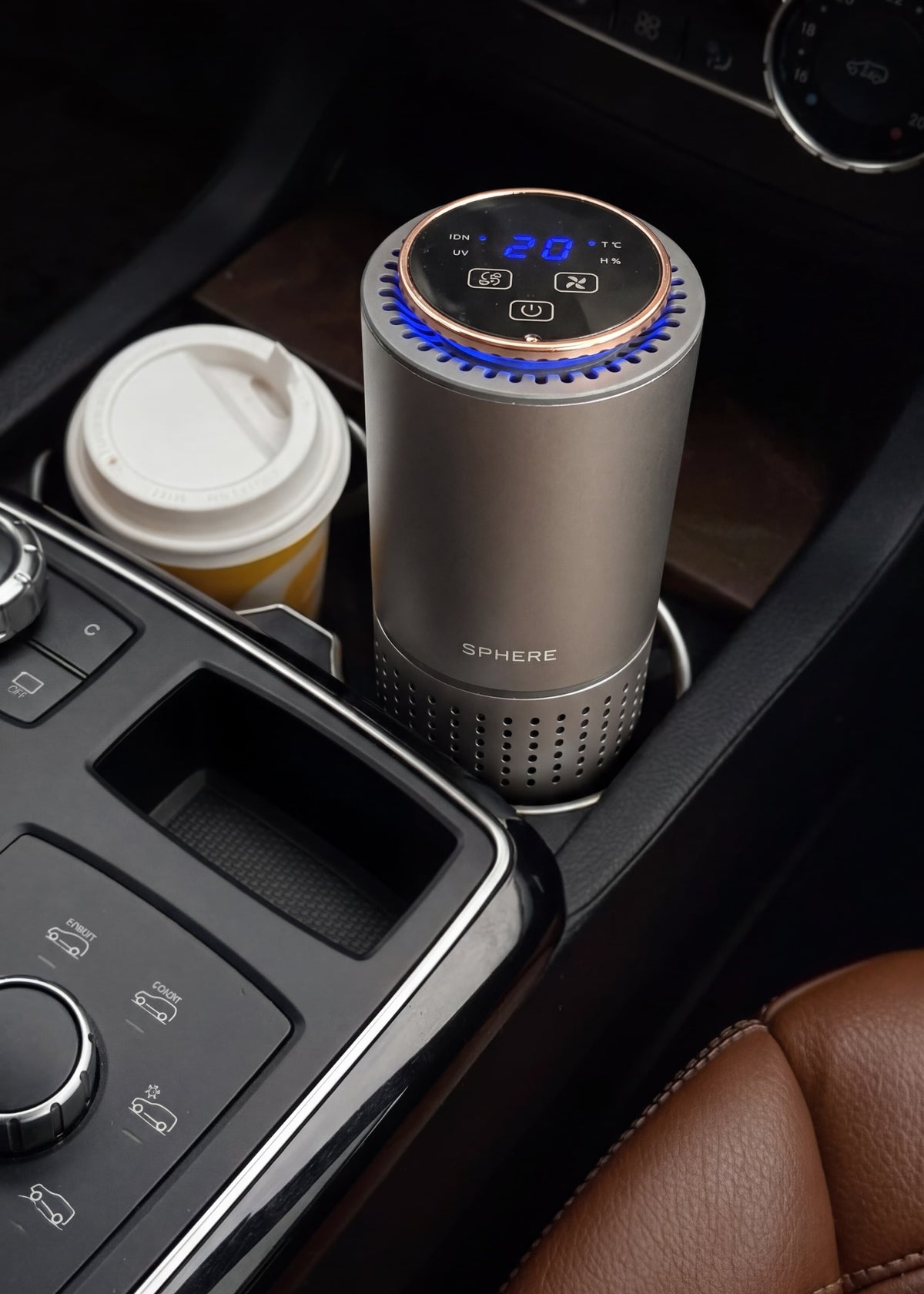 A silver cylindrical car air freshener diffuser with a digital display and blue LED light ring, placed in a cup holder of a car console next to a coffee cup.
