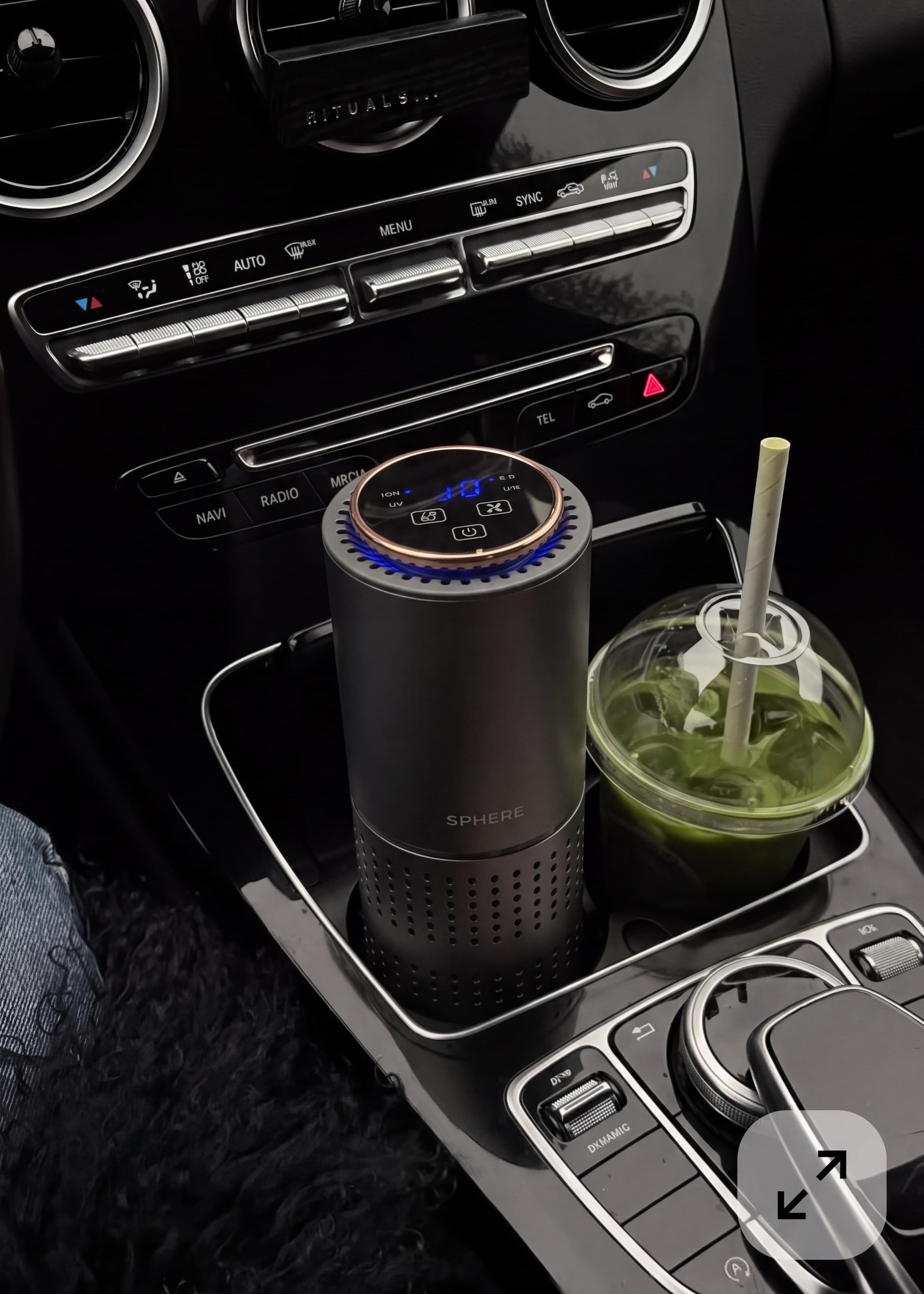 A cylindrical car air freshener diffuser with a digital blue LED display sits in the center console cup holder next to a green drink, inside a car interior.