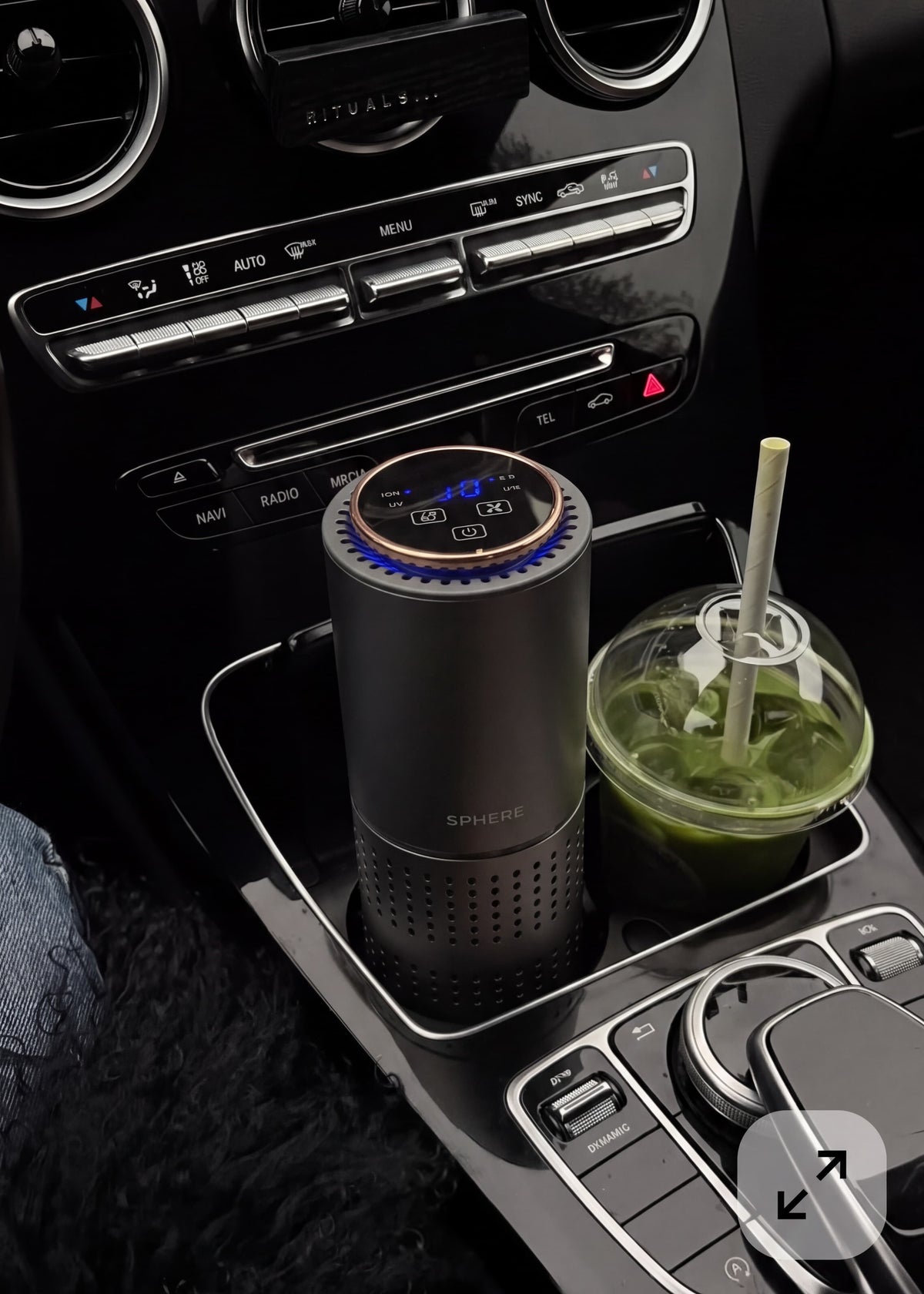 A cylindrical car air freshener diffuser with a digital blue LED display sits in the center console cup holder next to a green drink, inside a car interior.